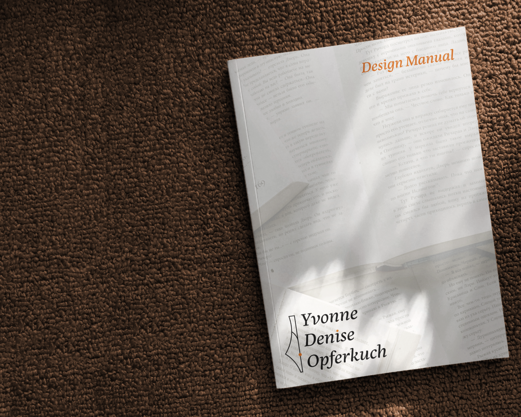 Design-Manual mit dem Namen „Yvonne Denise Opferkuch“ liegt auf einem braunen, grob gewebten Teppich; Sonnenlicht fällt durch ein Fenster und erzeugt weiche Licht- und Schattenmuster auf dem hellen Cover.