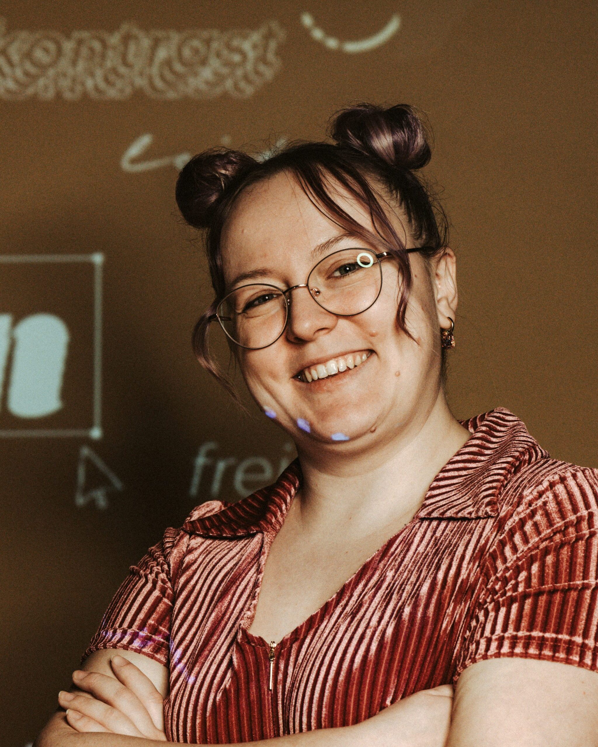 Porträt von Stranzi, der „Grafik-Magierin“ von Studio2: Eine Frau mit zwei Dutts und runder Brille lächelt direkt in die Kamera, die Arme locker verschränkt. Sie trägt ein rötlich schimmerndes, geripptes Oberteil und kleine Ohrringe. Auf den warmen Hintergrund und teilweise auf ihr Gesicht sind grafische Elemente und Schrift projiziert, die dem Bild eine kreative, spielerische und digitale Atmosphäre verleihen.