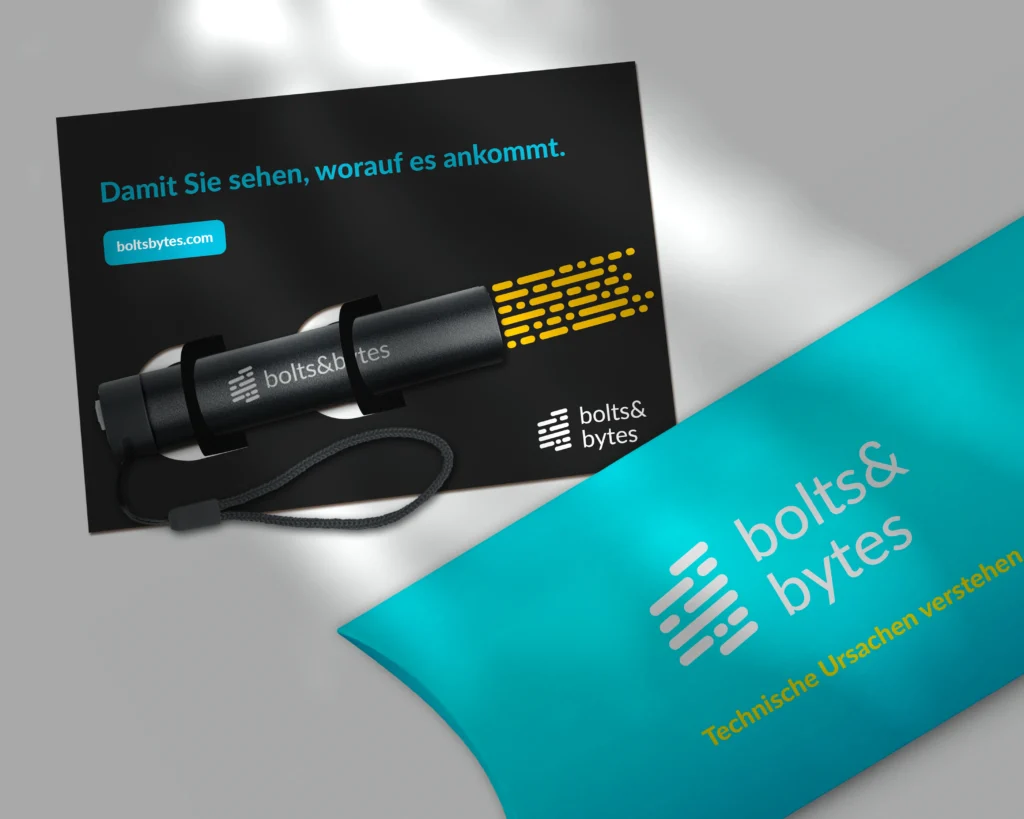 Türkisfarbene Kissenverpackung von bolts&bytes auf hellem Hintergrund, fotografiert in einer hochwertigen Produktinszenierung. Auf der Verpackung sind das bolts&bytes-Logo und der Schriftzug „Technische Ursachen verstehen“ zu sehen. Daneben liegt eine schwarze Postkarte mit dem Slogan „Damit Sie sehen, worauf es ankommt.“ sowie eine schwarze Taschenlampe mit Handschlaufe. Weiches Licht und dezente Schatten verleihen der Szene einen modernen, professionellen Look.