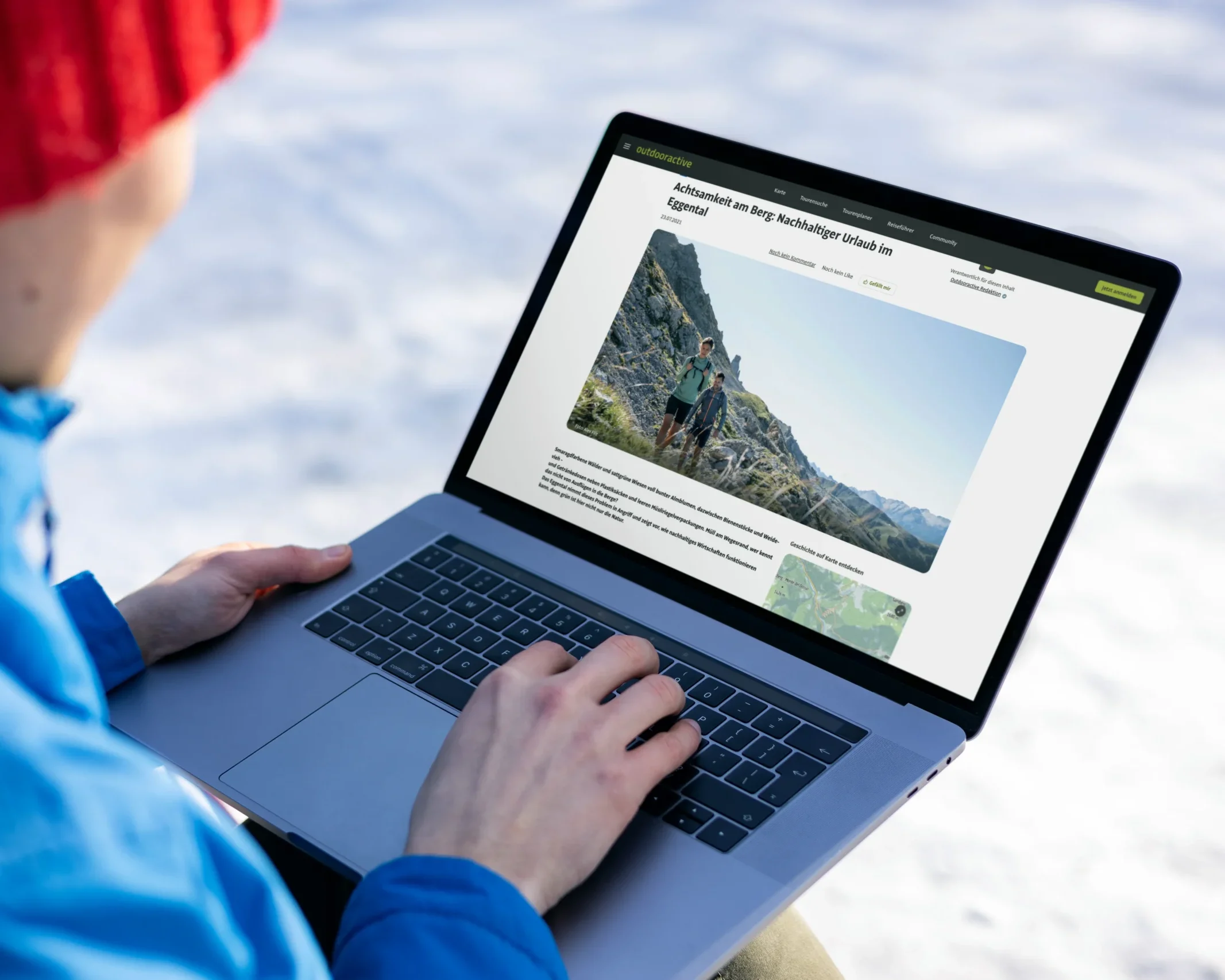 Person mit roter Mütze und blauer Outdoorjacke arbeitet draußen im Schnee an einem Laptop. Auf dem Bildschirm ist ein Artikel geschrieben von Sybille über nachhaltigen Urlaub und Achtsamkeit in den Bergen auf der Plattform Outdooractive geöffnet, mit großem Bergfoto und Wander:innen im Fokus.