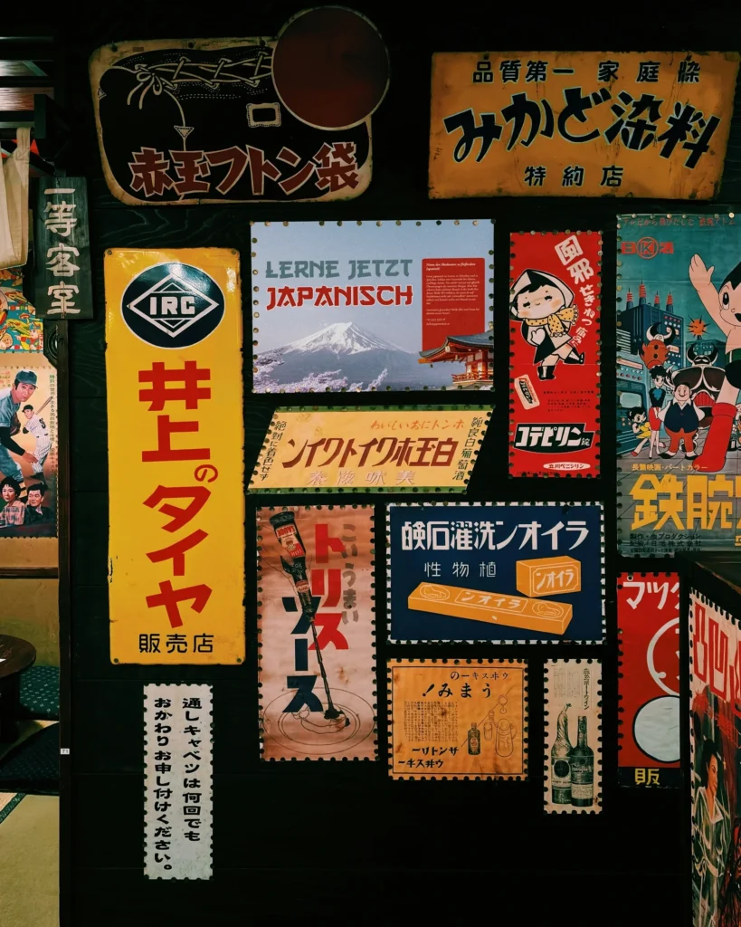 Wand mit historischen japanischen Werbeschildern und Plakaten, darunter ein Plakat mit dem Schriftzug „Lerne jetzt Japanisch“ und einer Abbildung des Fuji.