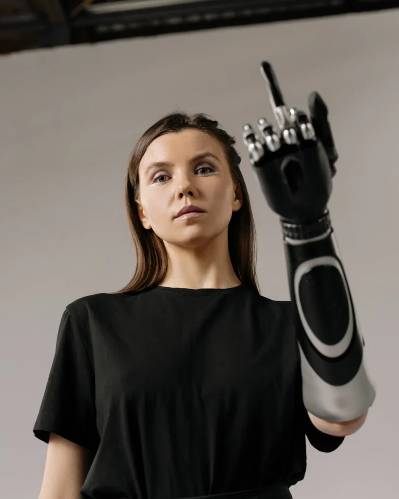 Frau in schwarzem Shirt hebt eine bionische Armprothese und zeigt damit den Mittelfinger vor grauem Hintergrund.