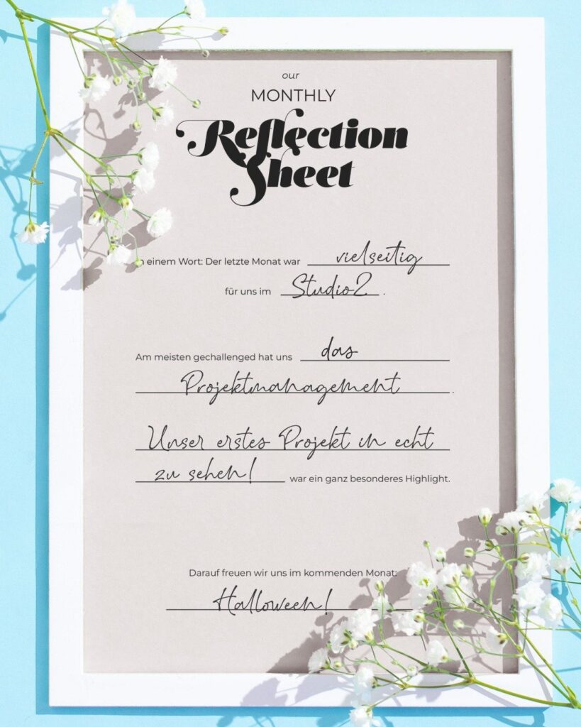 Monatliches Reflection Sheet von Studio2 mit handschriftlich ausgefülltem September-Rückblick: Der Monat war vielseitig, Projektmanagement war die größte Herausforderung, das erste Projekt in echt ein besonderes Highlight und Halloween die Vorfreude für den kommenden Monat. Das Blatt liegt in einem weißen Rahmen und ist mit weißen Blumen dekoriert.